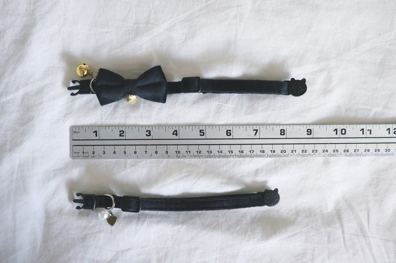 May include: Two black cat collars with different charms. The top collar has a black bow tie with gold bells. The bottom collar has a small silver heart charm with a pearl.