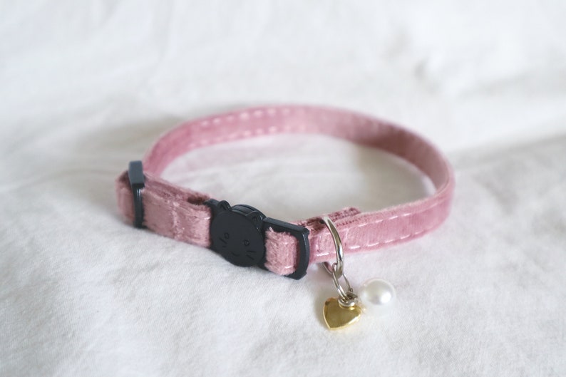May include: A pink velvet cat collar with a black cat-shaped buckle and a gold heart charm with a white pearl.