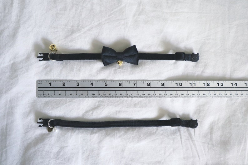 May include: A black cat collar with a black bow tie and a bell. The collar is adjustable and has a safety buckle. The collar is measured against a ruler with markings from 1 to 14.