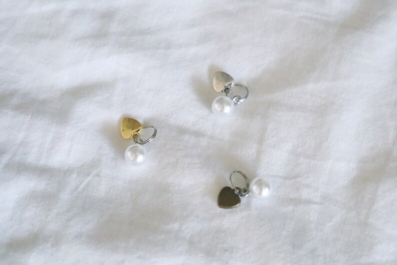 May include: Three pairs of heart-shaped earrings with pearl accents. The earrings are silver, gold, and black.