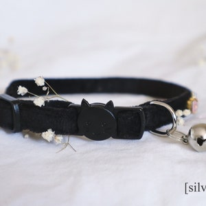 May include: Black velvet cat collar with a black plastic cat-shaped buckle and a silver bell. The collar has a small embroidered design with pink and white flowers.