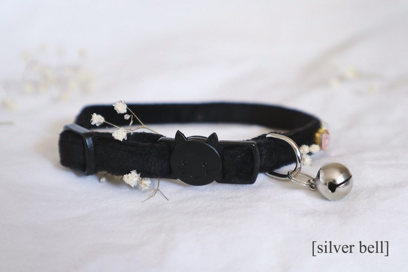 May include: Black velvet cat collar with a black plastic cat-shaped buckle and a silver bell. The collar has a small embroidered design with pink and white flowers.