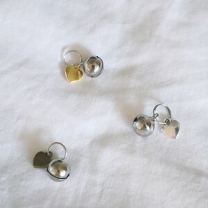 May include: Three silver bells with heart-shaped charms. The bells are round and have a shiny finish. The charms are attached to the bells with a small ring.