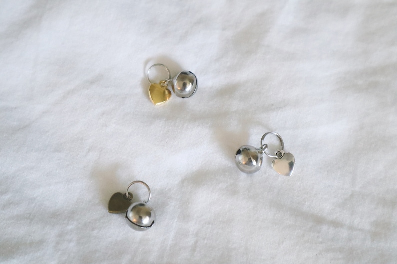 May include: Three silver bells with heart-shaped charms. The bells are round and have a shiny finish. The charms are attached to the bells with a small ring.
