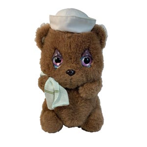 May include: A brown teddy bear toy wearing a white sailor hat and holding a light green handkerchief. The bear has large, iridescent eyes and a black nose. A vintage collectible item.