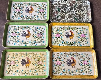 May include: A collection of six rectangular serving trays. Five trays feature a central rooster illustration surrounded by pink flowers and blue vines, with green or yellow borders. One tray displays a dense, dark floral pattern.