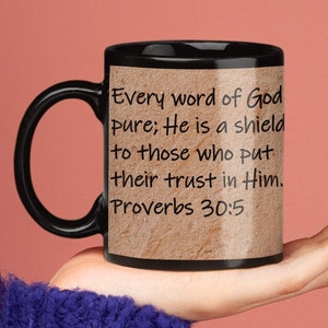 Puede incluir: Taza de café negra con fondo beige y el texto "Every word of God pure; He is a shield to those who put their trust in Him. Proverbs 30:5"