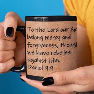 May include: Black coffee mug with a white rim and a beige background. The mug has a quote from the Bible, "To the Lord our God belong mercy and forgiveness, though we have rebelled against Him. Daniel 9:9" printed in black.