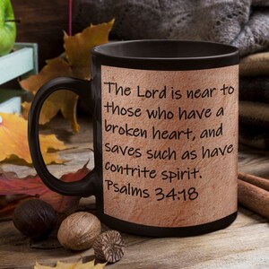 May include: Black coffee mug with a beige background featuring the text "The Lord is near to those who have a broken heart, and saves such as have contrite spirit. Psalms 34:18".