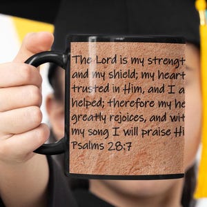 May include: Black ceramic mug with a black handle. The mug features the text "The Lord is my strength and my shield; my heart trusted in Him... Psalms 28:7" in black font on a textured, tan background.