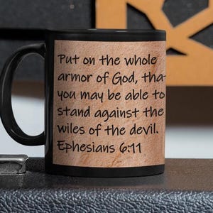 以下が含まれることがあります： 木目調のデザインが施された黒いコーヒーカップ。カップにはエフェソス人への手紙6章11節の言葉が書かれています。「悪魔の策略に立ち向かうために、神の完全なよろいを身につけなさい。」
