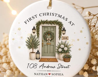 May include: A round white ceramic ornament featuring a printed illustration of a green front door with a wreath, potted plants, and lanterns. The text reads FIRST CHRISTMAS AT, 108 Andrew Street, NATHAN heart SOPHIA, and 2026. It hangs by a gold cord.