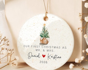 May include: A round, speckled white ceramic ornament hanging from a gold string. It features a watercolor illustration of a small Christmas tree in a basket, with the text "OUR FIRST CHRISTMAS AS MR. & MRS." above the names "Daniel" and "Kristine" with a red heart, and the year "2026" at the bottom.