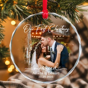 May include: A clear, faceted glass Christmas ornament with a red ribbon. The ornament features a photo of a couple and the text "Our First Christmas" and "married". The names "Amelia & Edward" and the date "11.25.2025" are also visible.