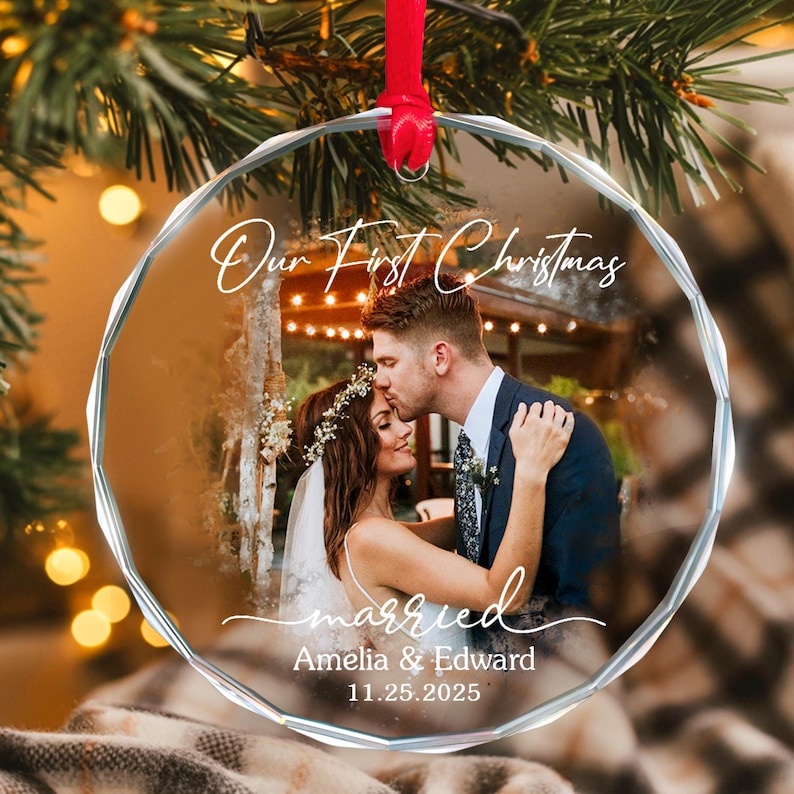 May include: A clear, faceted glass Christmas ornament with a red ribbon. The ornament features a photo of a couple and the text "Our First Christmas" and "married". The names "Amelia & Edward" and the date "11.25.2025" are also visible.