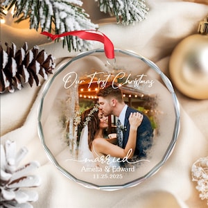 May include: A clear, faceted glass Christmas ornament with a red ribbon. The ornament features a photo of a couple kissing, with the words "Our First Christmas" and "married" written on it. The names Amelia & Edward and the date 11.25.2025 are also visible.