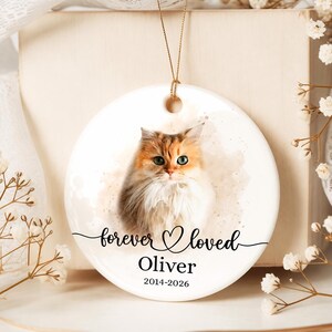 May include: A round white ceramic ornament featuring a watercolour-style portrait of a long-haired calico cat. The text "forever loved" is printed in black script above the name "Oliver" and the years "2014-2026" in a simple black serif font.