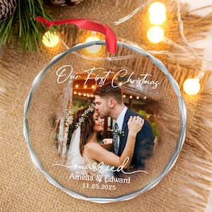 May include: A clear, round Christmas ornament with a photo of a couple kissing. The ornament has the words "Our First Christmas" and "married" written on it, along with the names "Amelia & Edward" and the date "11.25.2025". A red ribbon is attached.