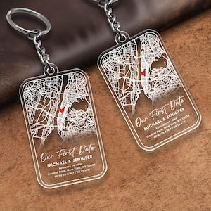 May include: Two clear acrylic keychains with silver chains. Each keychain features a white map design with a red heart and the text "Our First Date", along with names, a date, and coordinates.