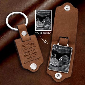 May include: A brown leather keychain with a silver key ring and a rectangular tag. The tag has the text "To Daddy. Now you can carry me too. Can't wait to meet you! Love from bump!" and a silver snap. The keychain also includes a photo of a baby ultrasound.