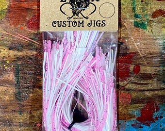 Könnte beinhalten: Eine durchsichtige Plastiktüte gefüllt mit pinken und weißen Glitzer-Angeljig-Strängen. Das Etikett lautet "English Angling E.A.A. Custom Jigs".