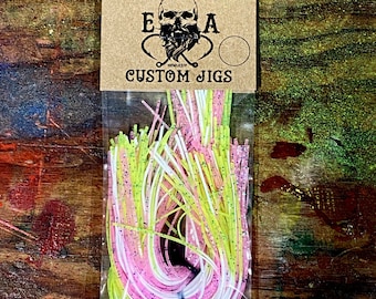 Könnte beinhalten: Eine Packung mit pinkfarbenen, weißen und gelben, maßgefertigten Jigging-Röcken mit dem Text "ENGLISH ANGLING E.A.A. CUSTOM JIGS" auf der Vorderseite.