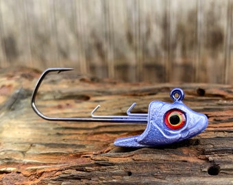 Könnte beinhalten: Ein blauer Angel-Jigkopf mit einem großen, rot-gelben Auge und einem gebogenen Haken. Der Jigkopf ist wie ein Fischkopf geformt und für den Einsatz beim Angeln konzipiert. Der Hintergrund ist eine verwitterte Holzoberfläche.