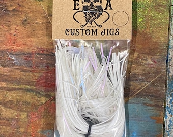 Könnte beinhalten: Ein transparenter Plastikbeutel gefüllt mit weißen, schillernden und rosafarbenen Angeljig-Strängen. Der Beutel trägt ein Etikett mit der Aufschrift "ENGLISH ANGLING E.E.A. CUSTOM JIGS".