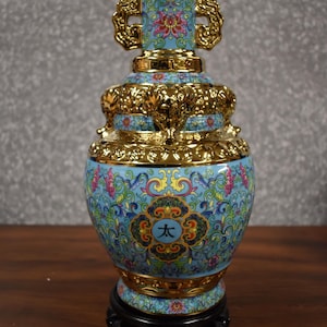 May include: A blue and gold Chinese porcelain vase with intricate floral designs and a gold trim. The vase has a round body and a tall, slender neck. The vase is decorated with a large, central flower and smaller flowers and leaves. The vase is sitting on a black wooden stand.