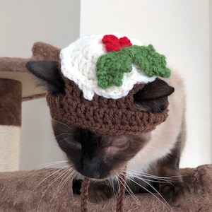 May include: A brown and white cat wearing a crocheted Christmas pudding hat with green holly and red berries. The cat is sitting on a brown cat tree. The text "CatsInHatsByMeg" and "@sia_josephine" is below the cat.