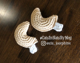 May include: Two crocheted cat ears in a light brown color with white tips. The ears are shaped like a crescent moon. The text #CatsInHatsByMeg and @sia_josephine is visible in the image.