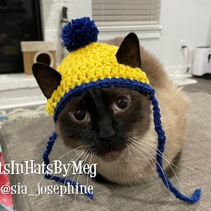 May include: A Siamese cat wearing a handmade yellow and blue crochet hat with a blue pom-pom and ear flaps. The hat is a vibrant accessory. The image includes the text "#CatsInHatsByMeg" and "@sia_josephine".