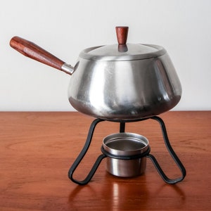 Peut inclure: Une fondue en argent avec une poignée en bois et un support en métal noir. La casserole a un couvercle avec un bouton en bois.