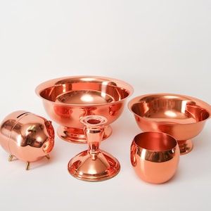 May include: A set of five copper-colored metal objects, including a bowl, a candlestick, a small cup, and a piggy bank. The objects are arranged on a white surface.