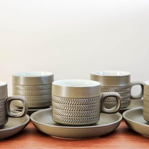 May include: A set of six ceramic coffee cups and saucers. The cups are olive green with a textured, woven pattern around the middle. The saucers are a matching olive green. The cups have a square handle and a white interior.