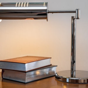May include: A chrome desk lamp with a cylindrical shade and adjustable arm, illuminating two hardcover books on a wooden surface.