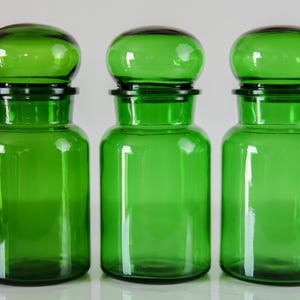 May include: Three matching apothecary jars made of translucent emerald green glass. Each jar features a cylindrical body with a wide mouth and a rounded, dome-shaped glass stopper that sits securely within the neck of the container.