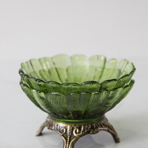 May include: A green glass bowl with a textured surface and a scalloped edge, resting on a decorative brass-toned metal stand with three legs. The bowl has a flower-like shape.