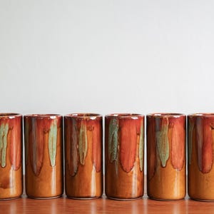 May include: A set of six cylindrical ceramic tumblers featuring a warm brown base glaze with vertical drips of deep red and sage green running from the rim. Each vessel has a smooth, glossy finish and a straight, uniform shape without handles.