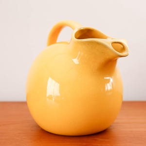 May include: A round, yellow ceramic pitcher with a handle and a spout. The pitcher has a glossy finish and sits on a wooden surface. The handle is arched, and the spout is designed for easy pouring. The pitcher is a decorative item.