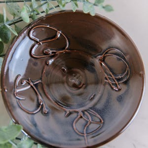 May include: A round, dark brown ceramic dish with a glossy finish. The surface features four raised, swirling line patterns embossed in a symmetrical arrangement around the center, which contains a small, circular indentation.