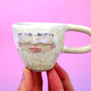 Op de afbeelding: Een keramische mok met een gezichtsontwerp. De mok is gebroken wit met een gespikkelde textuur en een gebogen handvat. Het gezicht heeft gesloten ogen, een neus en roze wangen en lippen. De mok wordt tegen een roze achtergrond gehouden.