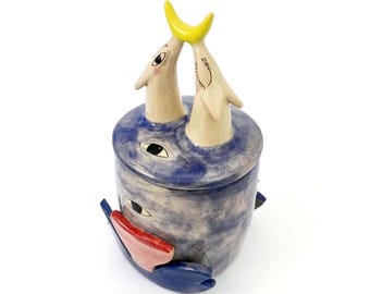 May include: A whimsical ceramic jar with a lid, featuring a blue and gray body with painted eyes and a red and blue accent. Two cream-colored figures with yellow crescent-shaped tops emerge from the lid, creating a unique decorative piece.