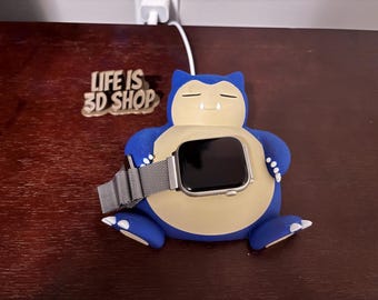 May include: A blue and tan Snorlax-shaped Apple Watch charging stand with an Apple Watch in place. The stand has the text "LIFE IS 3D SHOP" on a wooden plaque. The watch has a silver band.