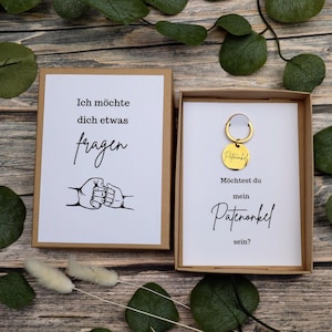 Könnte beinhalten: Ein Geschenkset mit einer Karte und einem Schlüsselanhänger. Die Karte lautet "Ich möchte dich etwas fragen" mit einer Grafik von Händen, die sich umarmen. Der Schlüsselanhänger ist goldfarben und trägt das Wort "Patenonkel". Die zweite Karte lautet "Möchtest du mein Patenonkel sein?"
