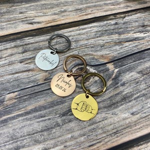 Puede incluir: Tres llaveros personalizados con etiquetas redondas de metal. Un llavero plateado tiene la palabra "Patenkind" grabada. Un llavero de oro rosa tiene el nombre "Nicolas" y la fecha "03.05.26". Un llavero dorado presenta un dise&ntilde;o de choque de pu&ntilde;os.