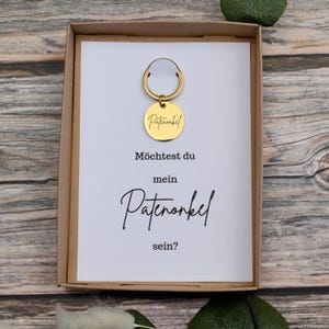 Puede incluir: Un llavero dorado con una etiqueta redonda dentro de una caja de regalo. La etiqueta tiene grabada la palabra "Patenonkel". La tarjeta dentro de la caja tiene el texto "M&ouml;chtest du mein Patenonkel sein?"