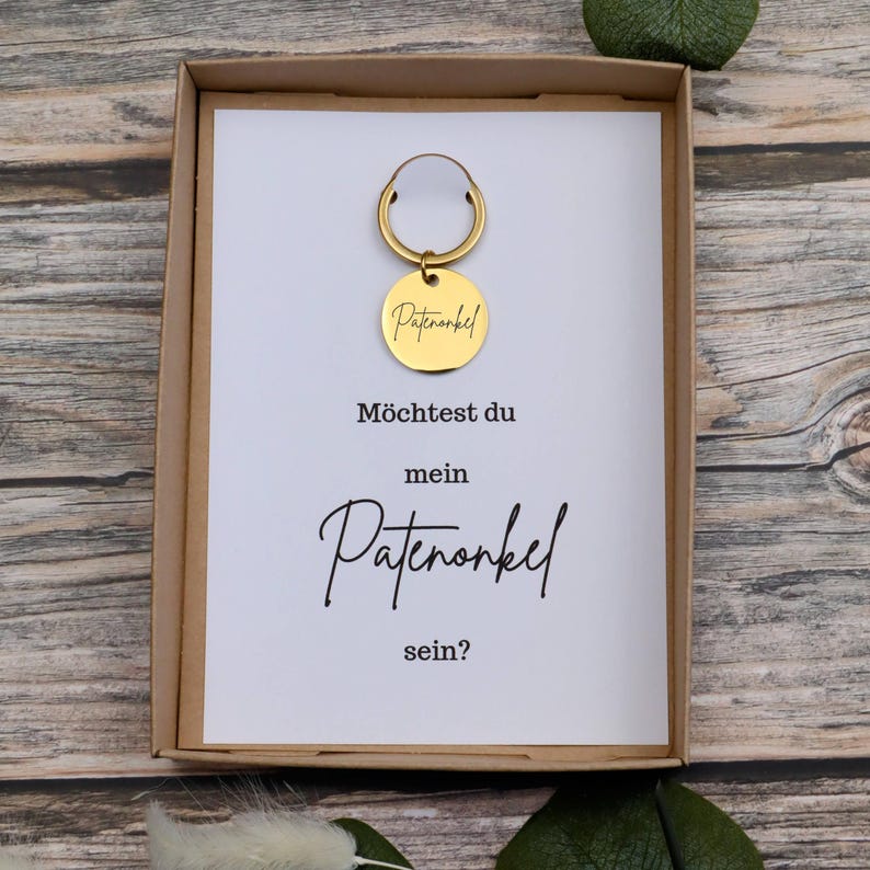 Puede incluir: Un llavero dorado con una etiqueta redonda dentro de una caja de regalo. La etiqueta tiene grabada la palabra "Patenonkel". La tarjeta dentro de la caja tiene el texto "M&ouml;chtest du mein Patenonkel sein?"