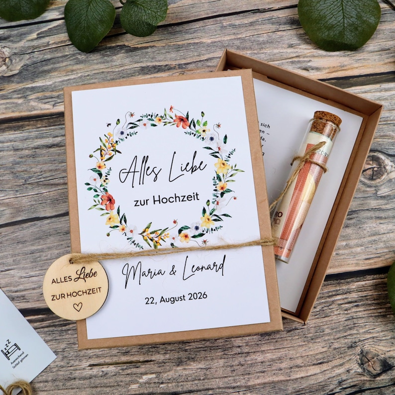 May include: A rectangular cardboard gift box containing a white card with a floral wreath illustration, the text Alles Liebe zur Hochzeit, the names Maria & Leonard, and the date 22, August 2026. A glass vial with a cork stopper sits inside the box.