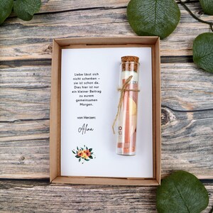 May include: A brown cardboard gift box containing a white card with German text and a small floral illustration. A clear glass vial with a cork stopper is tied to the card with twine, containing a rolled-up banknote inside the glass container.